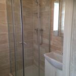 A corner shower cubicle with glass doors in a bathroom with beige, bathroom tiling.