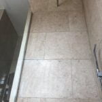 A view of a bathroom tiling area with beige tiles, featuring a shower head and a glass partition.
