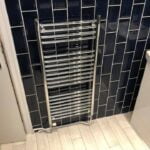 A towel radiator mounted on a wall with dark blue porcelain tiling, above white tiled flooring.
