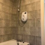 An electric shower unit mounted above a bathtub in a bathroom with mosaic tiling on the walls.