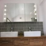 Modern bathroom vanity with two vessel sinks, a wooden cabinet, and a mirror with side lights set against porcelain tiling.