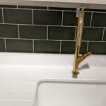 A gold faucet over a white sink against a green tiled bathroom backsplash.