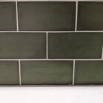 A bathroom wall with green porcelain rectangular tiles above a white baseboard.