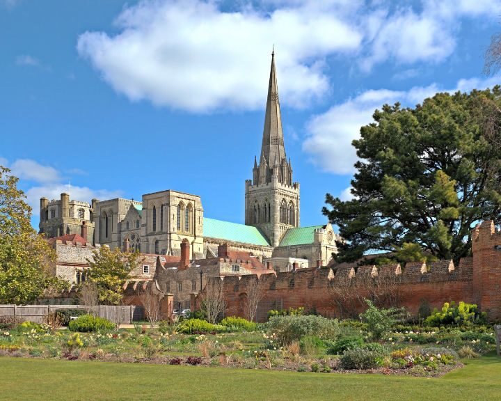 A historic cathedral with a tall spire, featuring mosaic tiling, rises above an enclosed garden under a clear blue sky.