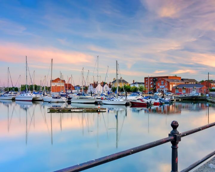 A serene marina at dusk with moored sailboats and reflections on the water, under a pastel sky with natural stone tiling along the waterfront.