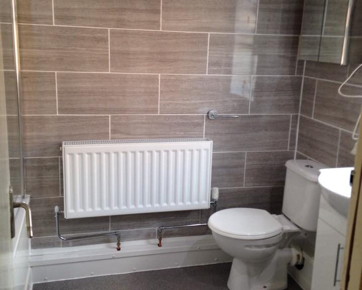 A modern bathroom with beige natural stone tiling, a white toilet, radiator, and gray flooring.