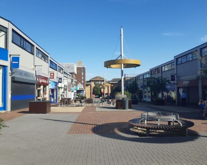 A pedestrian square with shops, a canopy structure, and seating under a clear blue sky features porcelain tiling.